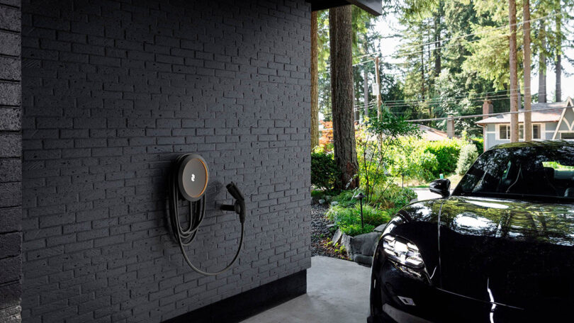 Cove EV charging system wall mounted onto a black brick carport with black EV parked to the right with forested residential neighborhood in the background.