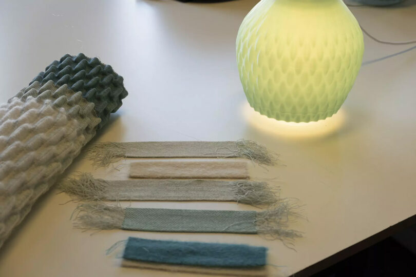 Five woven textile swatches and a roll of Aleksandra Gaca's woven textile sample set across a table next to an illuminated Philips Droplet Three Pendant light.
