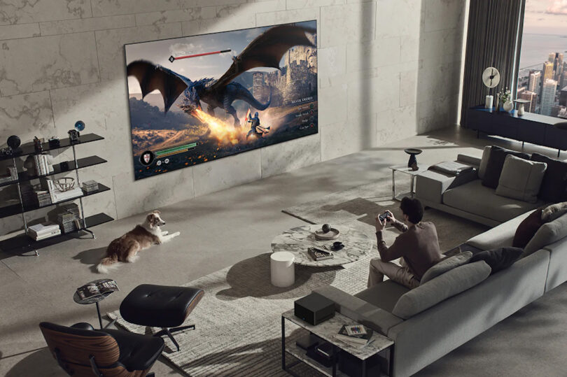 Overhead view of modern loft space with man seated on a gray modern sofa playing fantasy fiction video game on a wall-mounted 97-inch OLED television with Australian Shepherd dog laying down nearby.