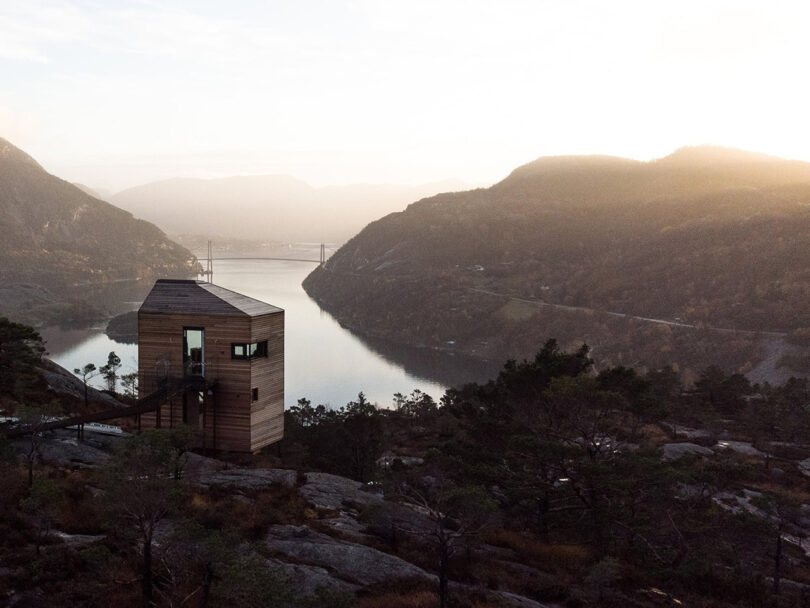 Silhouette of one of the The Bolder Lodges cabin during a hazy warmly illuminating sunrise with a bridge spanning the fjord in the distant background and the mountains reflected across the water.