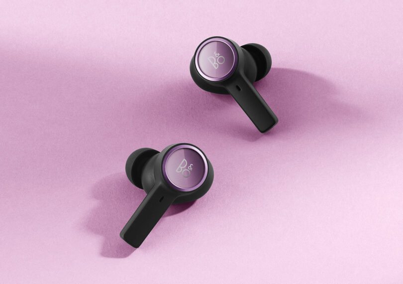 Two Beoplay EX Lilac Purple earbuds set on pink surface, with "B&O" logo subtly printed across each earbud with round Lilac circle.