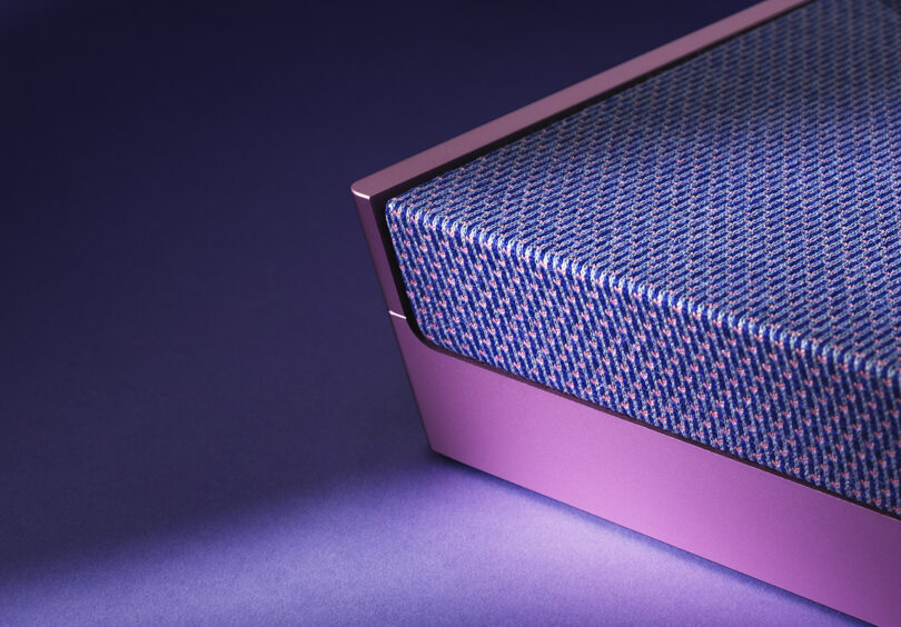 Detail corner of Fall 2023 Atelier Editions Beosound Level wireless speaker in Lilac Twilight color staged against a purple floor and background with shadows cast across it showing where the aluminum pink frame meets the speaker fabric front.