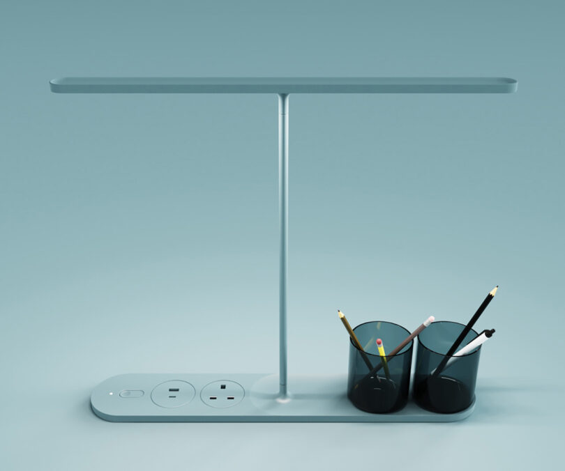 Current LED desk lamp set on light blue surface with two small storage cups filled with pens and pencils. The left side of lamp base includes a USB A and USB C ports, and a European compatible power outlet near the lamp's on/off switch.