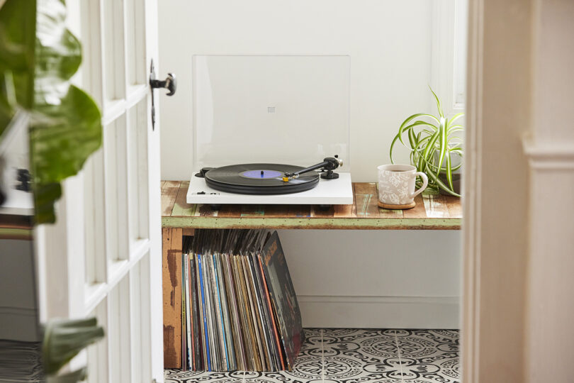 White ORBIT turntable with lid open and record on platter with tone arm playing record, placed on rustic wood bench with coffee mug and house plant to the left, and selection of records stored underneath across ornate black and white tile floor. A white window pane door is opened in the foreground.