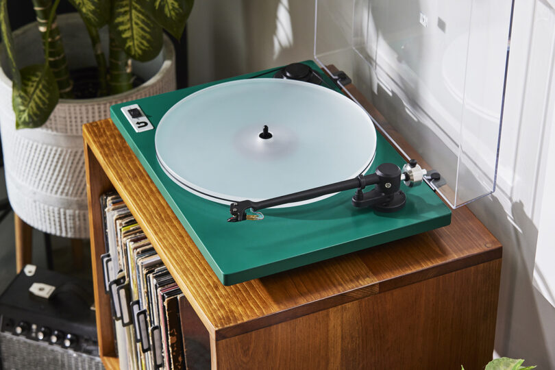 Angled view of ORBIT turntable with green plinth, clear acrylic platter, and cover open placed on top of a small vinyl record storage shelf with potted houseplant in the background. 