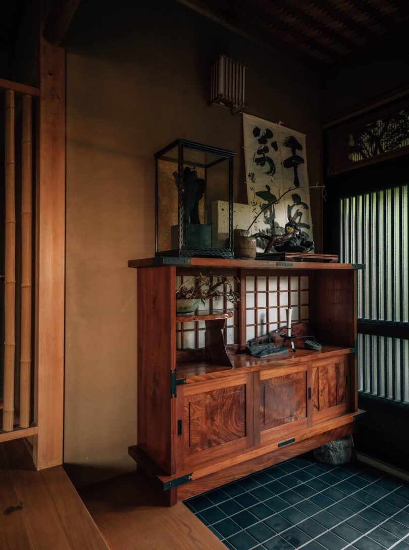 Entryway cabinet decorated with traditional Japanese artwork, scrolls, and small objects of decor set across black tile floor, sliding rice doors to the right and bamboo partition to the left.