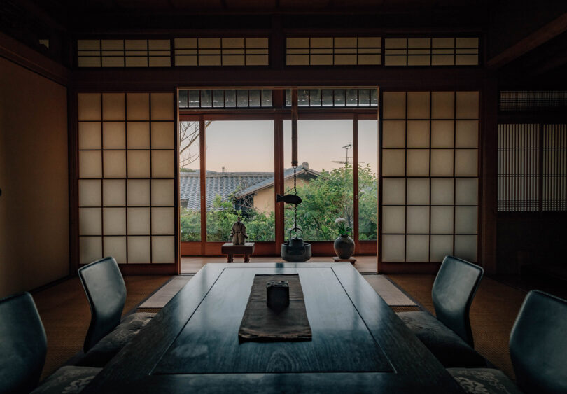 Dimly sunset-lit room with floor seating and center table for four people. In the background can be seen a nearby Japanese home's roof alongside a hanging tea kettle through two large rice paper screen doors.