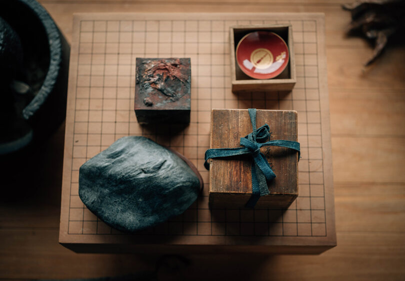 Overhead shot of traditional Japanese wood boxes, a large stone, and small lacquer bowl all set on top of a Go boardgame board.