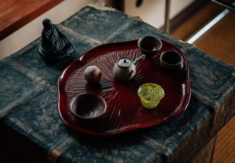 Red lotus leaf shaped lacquer tray with small cups and tea ceremony preparation items set across old weathered side table with small Buddha sculpture nearby.