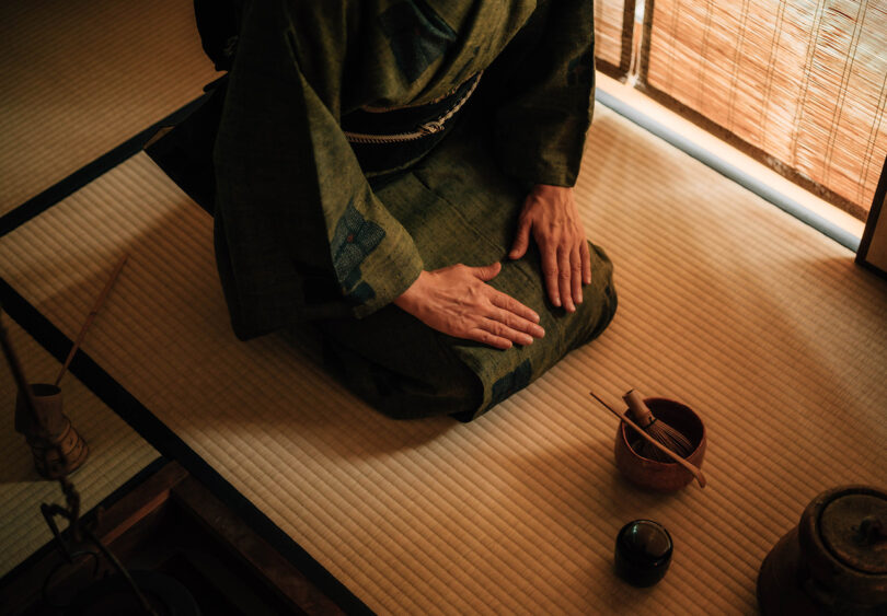 Japanese woman in traditional kimono kneeling with traditional Japanese tea ceremony bowl, whisk and scoop in front of her on tatami mat.