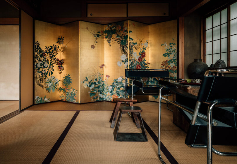 Golden gilded folding room screen decorated in traditional Japanese flower painting in background, with Wassily armchairs and small side stools near the front of the photo with tatami flooring throughout.