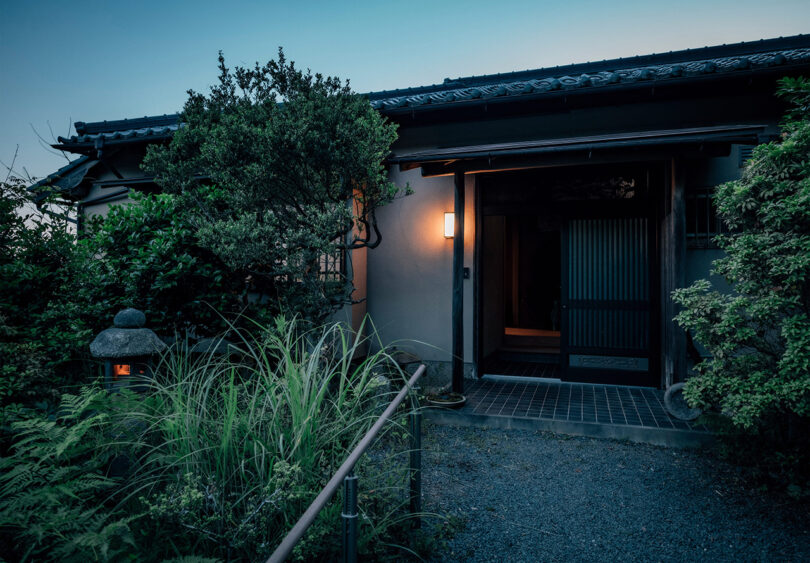 Front view of the home's small tea garden entry lined with gravel, surrounded by lush traditional Japanese landscaping and tile roof, gently illuminated at dusk by a wall mounted light and small Japanese stone lantern.