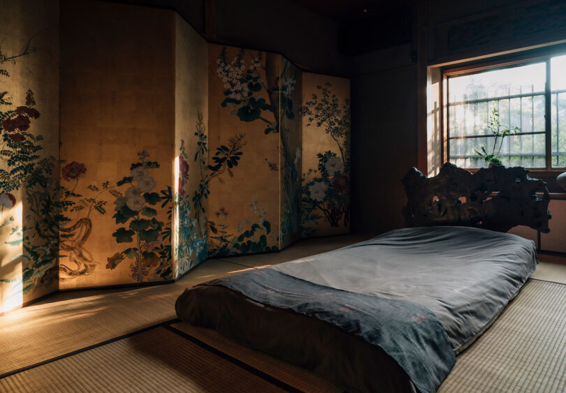 Traditional Japanese futon with ornate reclaimed wood headboard across tatami floors with wall-sized gilded and art decorated folded room screen.