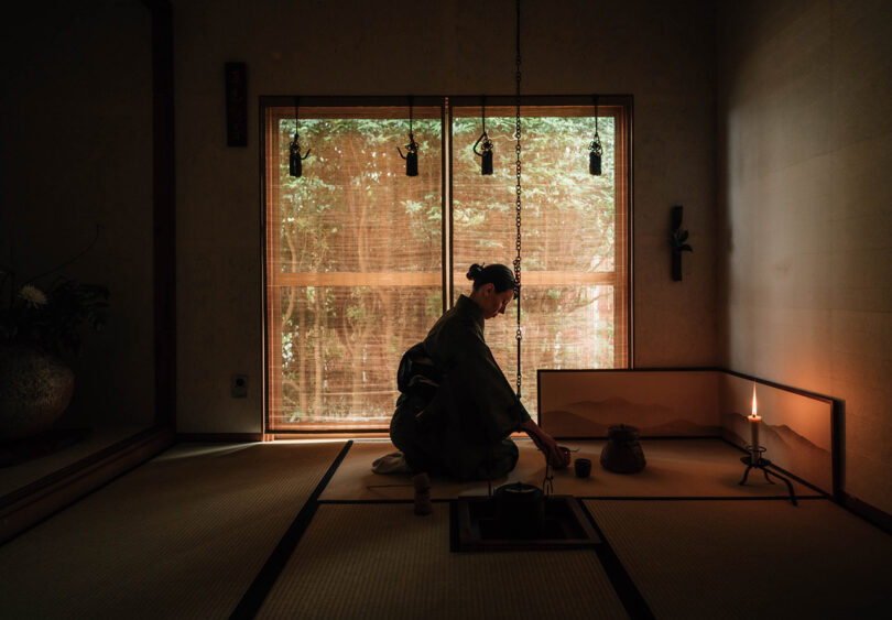 Japanese woman in traditional kimono kneeling with traditional Japanese tea ceremony bowl, whisk and scoop in front of her on tatami mat and lone candle to the right dimly illuminating the room's corner. Partially shade covered view of surrounding garden is visible.