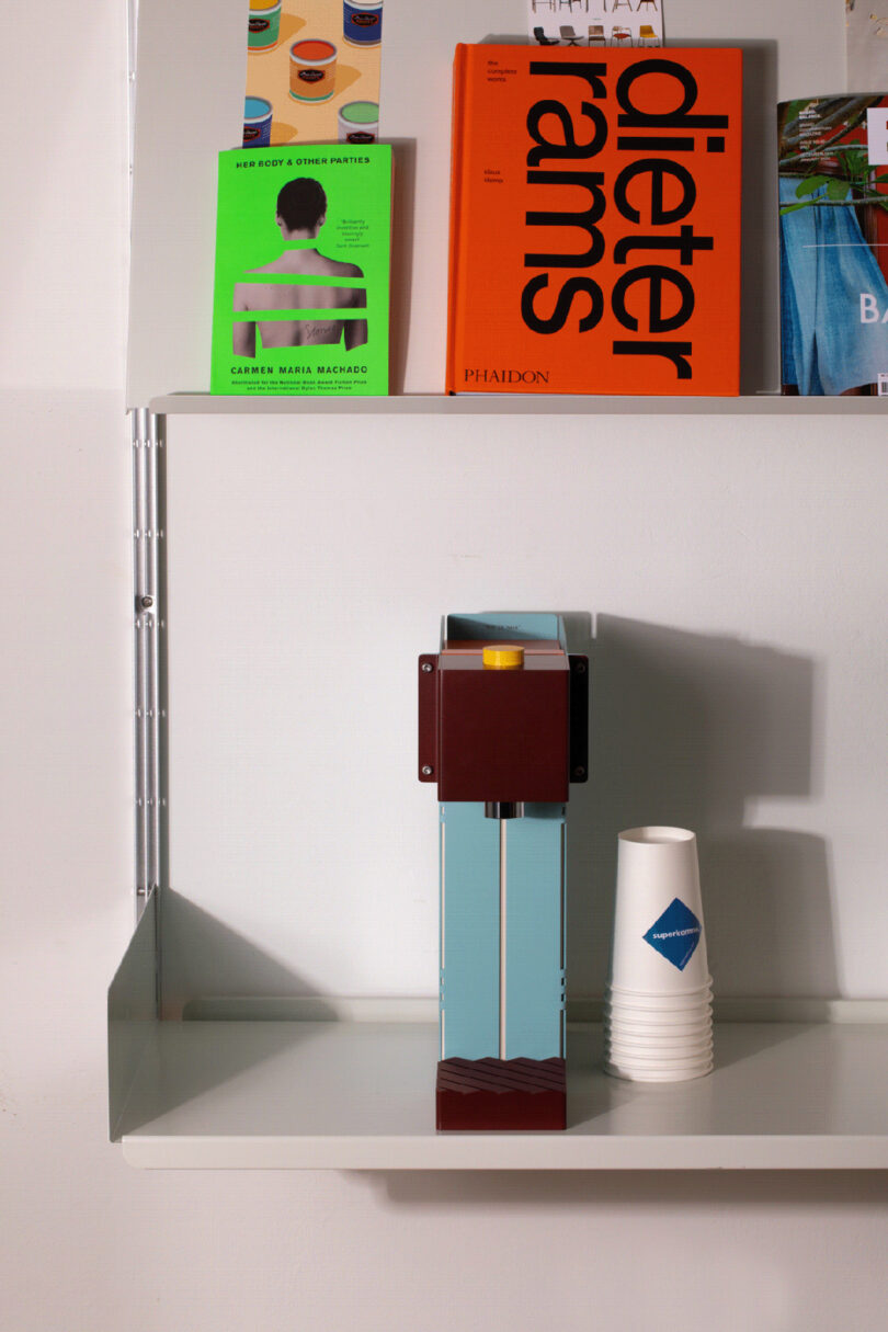 Colorful slim folded metal construction water purifier appliance viewed from the front in blue, brown and yellow on white floating shelf. A small group of nesting paper cups sit to the right of the Conecto, with a Dieter Rams book and other books situated above it on another display shelf.
