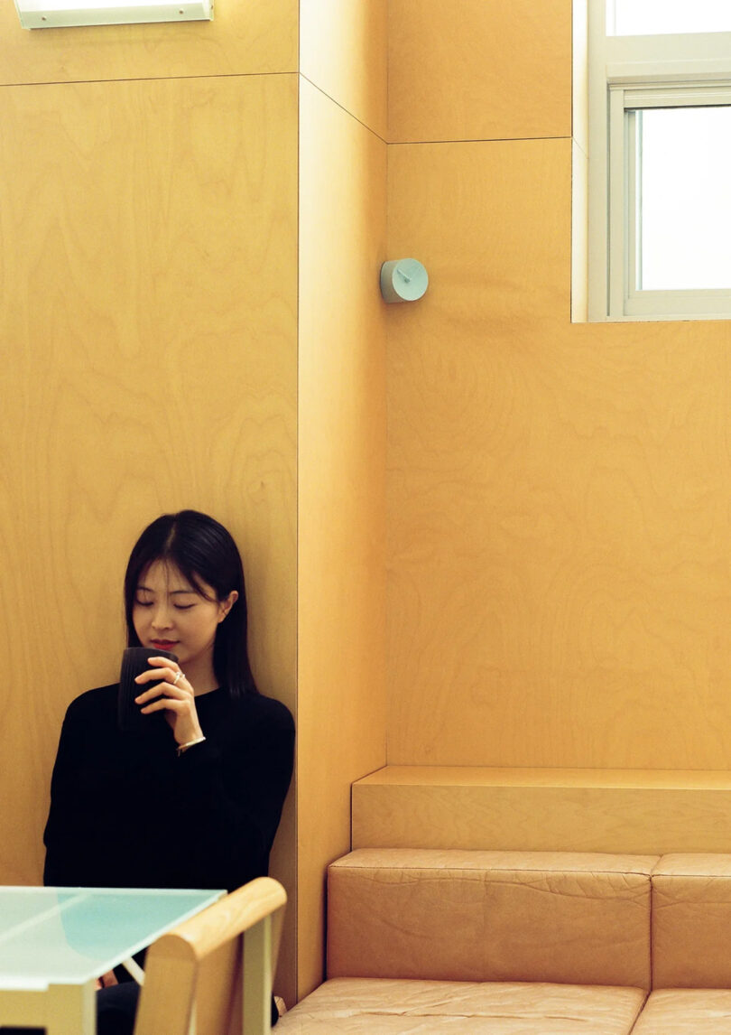 Young long haired Korean woman seated leaning against a plywood panel wall with small mug in hand and small light green analog clock installed into a corner just behind and above her left shoulder.