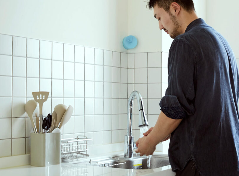 Young Korean man washing glasses at the sink with a light blue analog clock wedged into the corner above the kitchen backsplash white grid tiles.