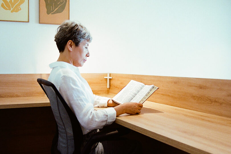 Person studiously reading the Bible at a corner wood desk, with small white cross in the corner with two modern framed prints partially in view.