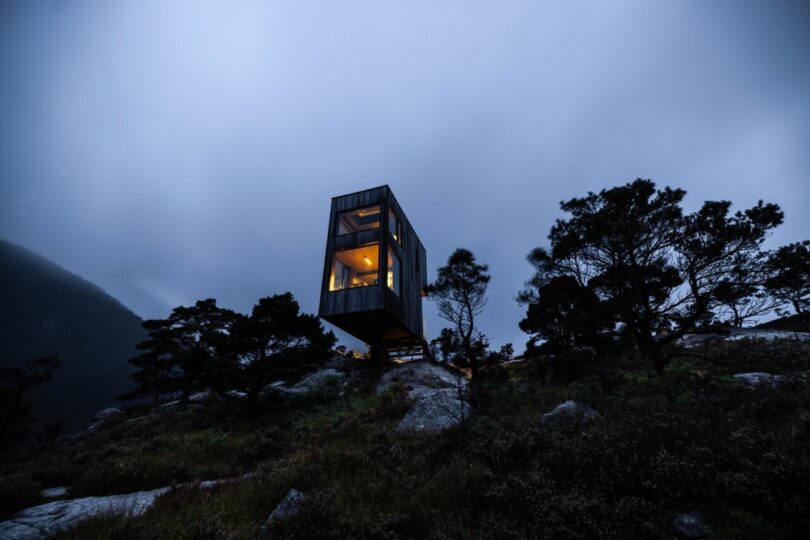 View of the of the two smaller 22 square meters SkyLodges overhanging the mountainside, captured during a dramatically darkened cloudy sky with the interior of the cabin warmly illuminated.