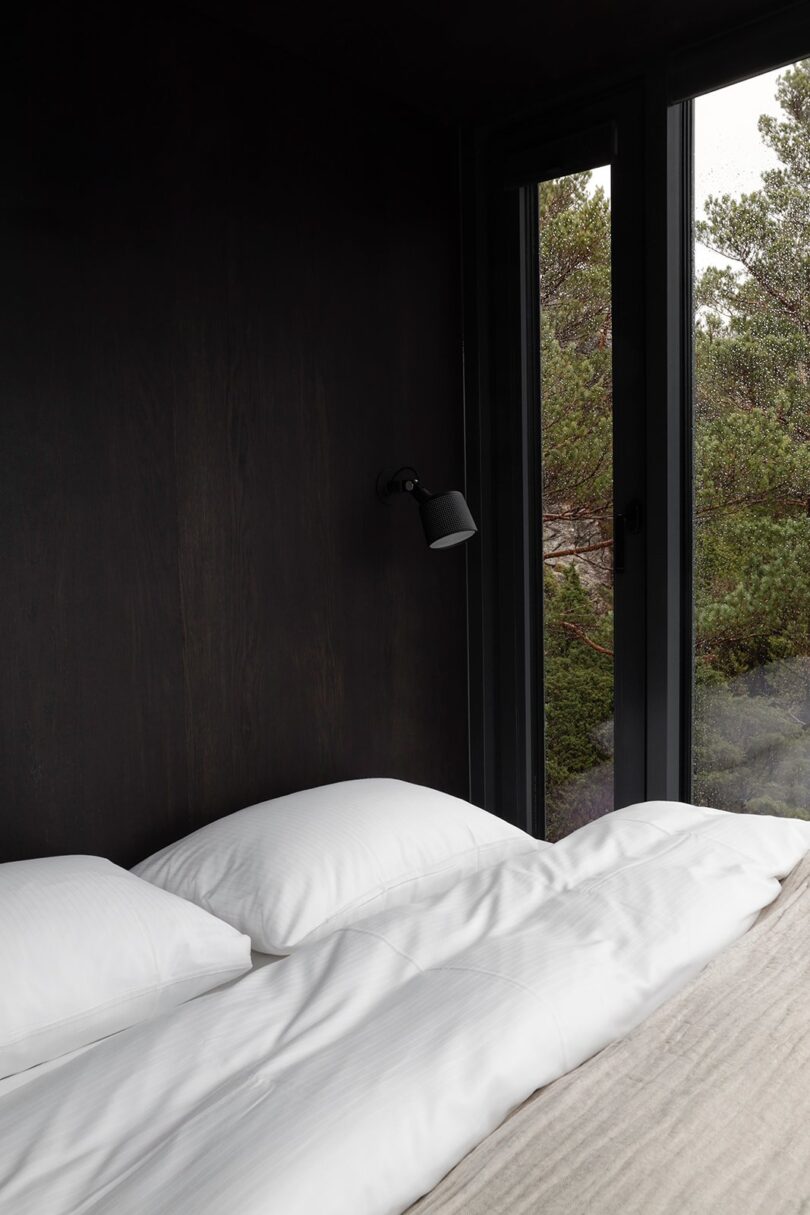 Dark cozy bedroom with white linens and view of surrounding forest through side windows. Small black sconce to the right of the bed.