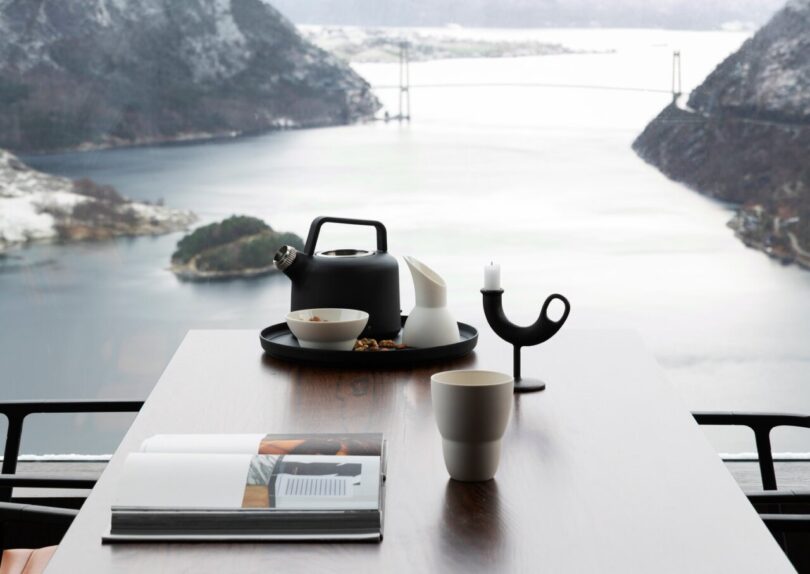 Dining table with Scandinavian modernist design tea kettle, candle holder, mug, creamer and small bowl with two dining chairs face to face with majestic view of the Norwegian fjord waters below.