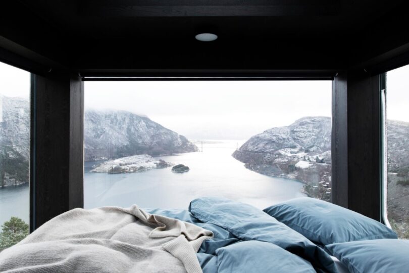 A majestic panoramic view of the nearby fjord located below the series of cabins from the glass facade covered bedroom, with light blue bedding and off-white blanket.