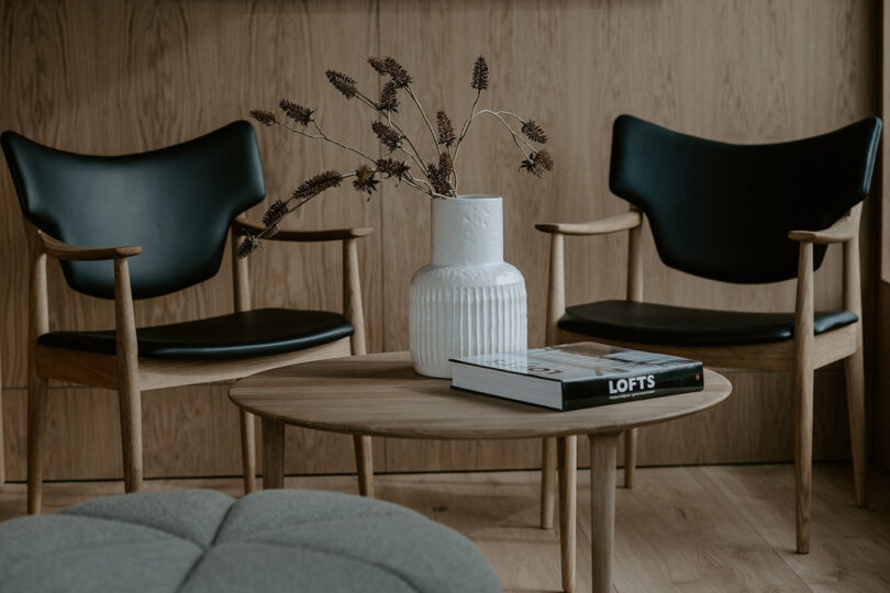 Two Scandinavian modern armchairs and small coffee table with white ceramic vase planter with small arrangement of dried flowers and "LOFTS" book on the surface; gray soft fabric pouf in the foreground.