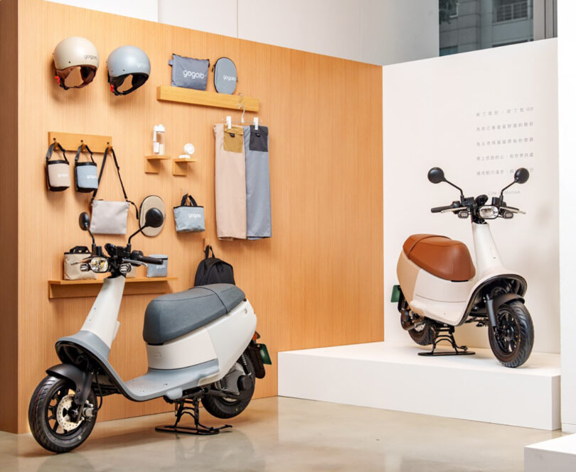 Two Muji Gogoro Smartscooter parked in a showroom with Muji styled accessories displayed on the wall, including a pair of helmets, bags, water bottle, and small personal fan.