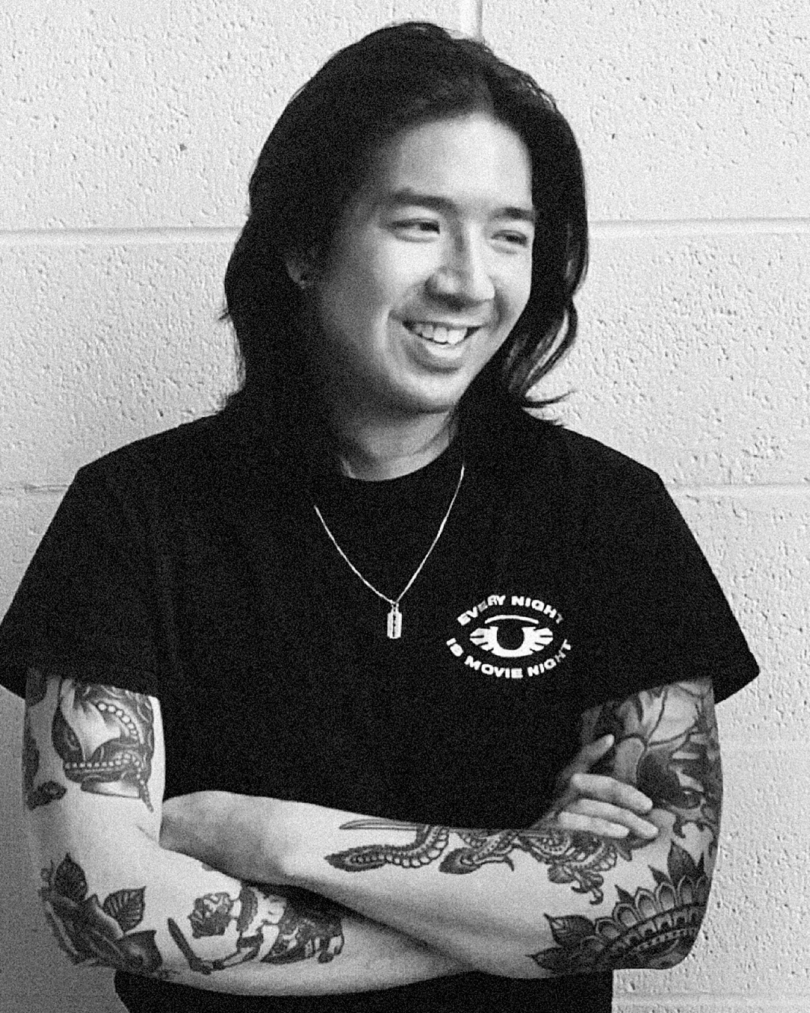 black and white image of a man with shoulder-length dark hair wearing a black t-shirt with his arms crossed and smiling