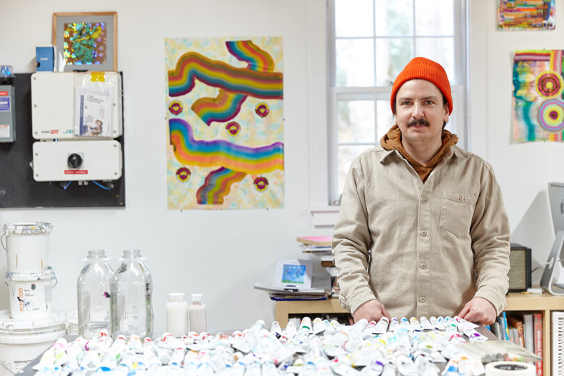 light-skinned man with a dar mustache wearing an orange hat and a khaki colored jacket while standing in a painting studio
