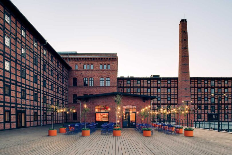 evening view of large brick industrial building with cafe on lower level with outdoor space filled with blue chairs and tables