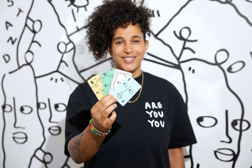 brown-skinned woman with pulled up curly black hair wearing a black t-shirt reading ARE YOU YOU hold up a hand of colorful cards
