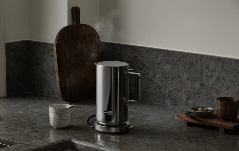 Stainless steel Aarke Kettle set kitchen counter top next to small ceramic tea drinking cup and small bowl of loose tea leaves. In the background is a oval shaped wooden cutting board with handle leaning up against the wall.