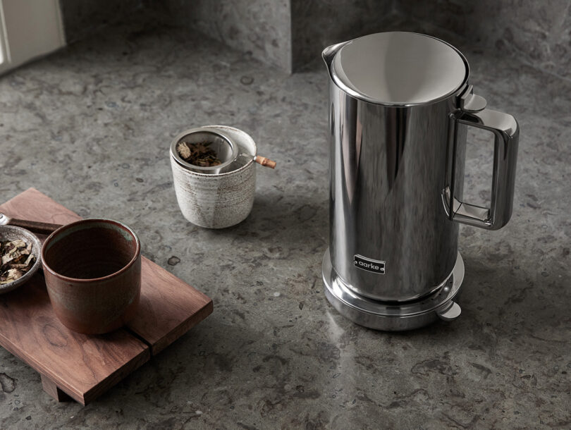 Stainless steel Aarke Kettle set on marble kitchen counter next to two small ceramic tea drinking cup and small bowl of loose tea leaves placed upon small wooden platform display.