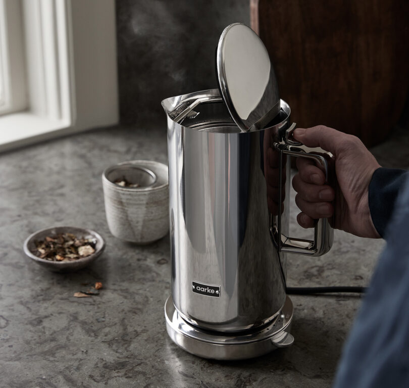 Man's hand pressing the lid button to open the Aarke Kettle's top lid. Nearby is a small bowl of loose tea and a small ceramic mug without handle with a tea leaf steeping mesh. A subtle plume of steam can be see escaping the electric kettle with its lid set vertically.