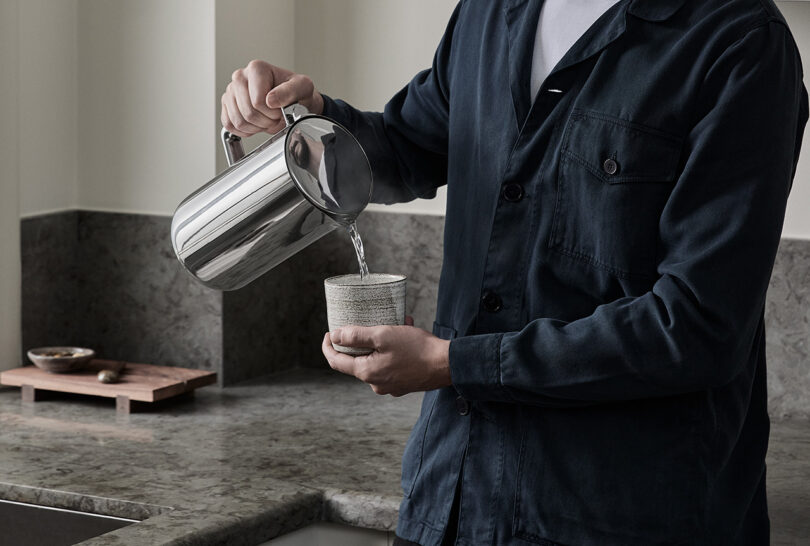 Man in dark blue button up shirt pouring heated water from Aarke Kettle into small ceramic tea drinking cup in a kitchen setting corner.