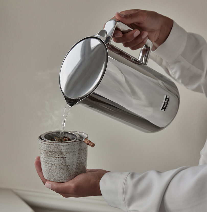 Person in long sleeve white shirt holding a small ceramic tea cup with loose leaf steeping mesh tool in their left hand while pouring hot water from the Aarke Kettle using their right hand. A small amount of steam can be seen coming from the cup.