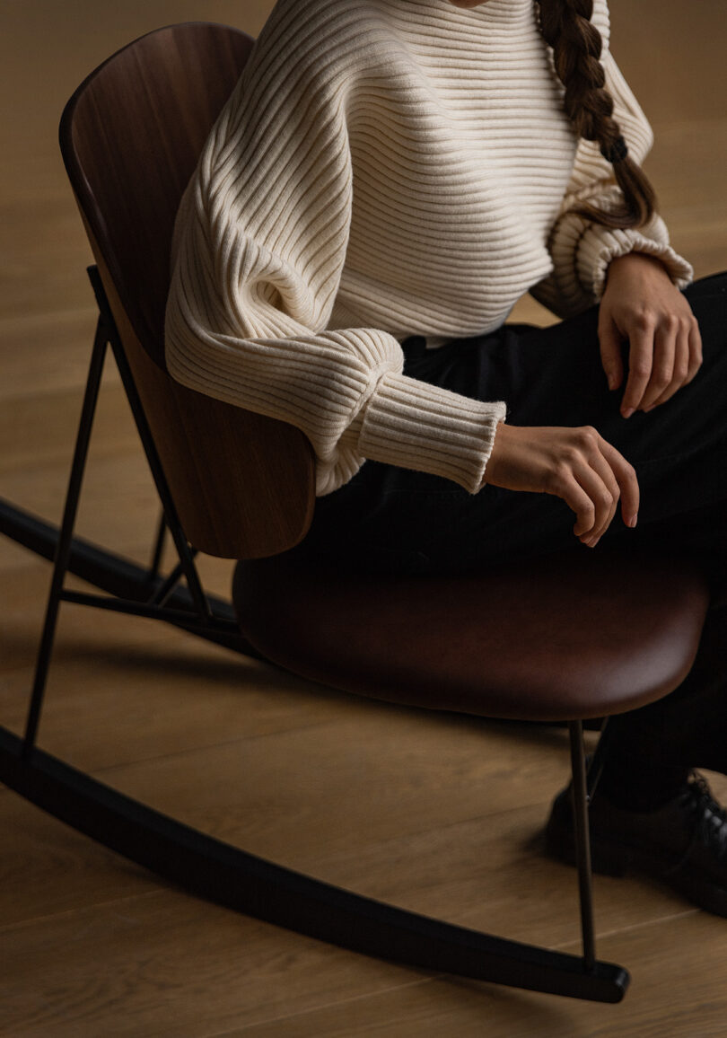 Woman with braided long hair in a ribbed off-white sweater leaning slightly forward while seated upon a Penguin Rocker Chair with leather upholstered seat and walnut back.