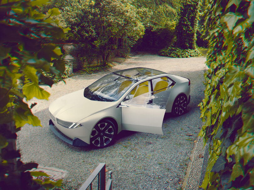 Angled overhead view of the BMW Vision Neue Klasse in white with yellow interior, with driver side door ajar. Car is parked on a gravel driveway surrounded by plants and trees.