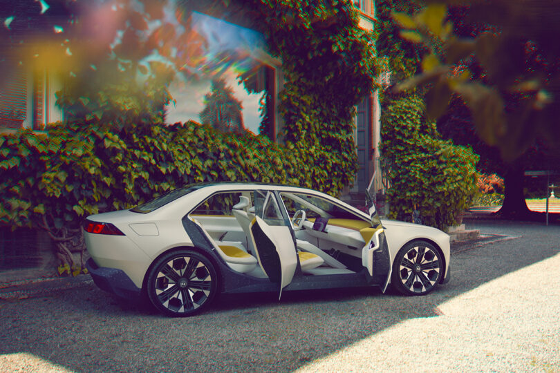 Side view of the BMW Vision Neue Klasse in white with yellow interior, with front passenger and rear passenger side doors ajar. Car is parked on a gravel driveway surrounded by plants and trees.