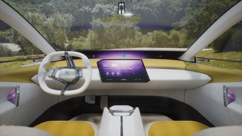 Interior cabin of the BMW Vision Neue Klasse concept with steering wheel on the left and angled infotainment display in the center. Interior is finished in yellow corduroy and white surfacing, with large windshield and side windows offering view of trees outside.