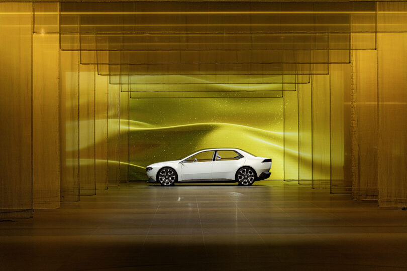 Distant view of BMW Vision Neue Klasse in white parked in an interior stage with a multitude of yellow semi-transparent planes with a backdrop of stars and undulating dune-like landscape behind the vehicle.