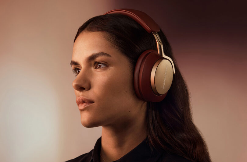 Beautiful woman with long brunette hair wearing the Bowers & Wilkins Px8 in Royal Burgundy Nappa leather finish with gold detailing staring to the left.