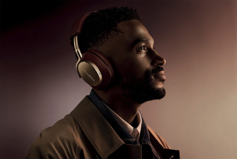 Handsome black man in slight shadow and light wearing a button up shirt and coat, staring to the right wearing Bowers & Wilkins Px8 in Royal Burgundy Nappa leather finish with gold detailing.