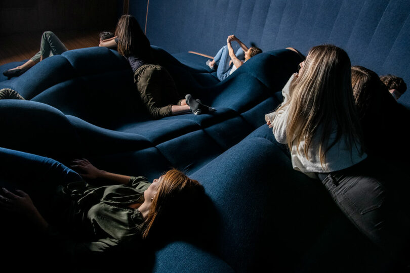 back angled view of home theater with wave-like dark blue seating with people sitting randomly view screen