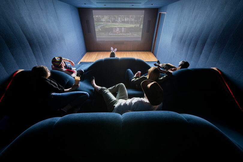 back view of home theater with wave-like dark blue seating with people sitting randomly in front of movie screen