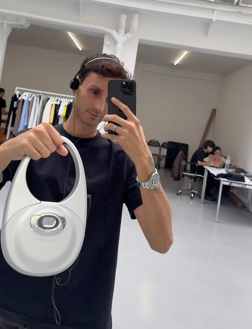 Coperni Creative Director & Co-Founder Sebastien Meyer holding up his brand's CD-PLAYER Swipe Bag with right hand, and photographing himself in the mirror with iPhone in left hand.