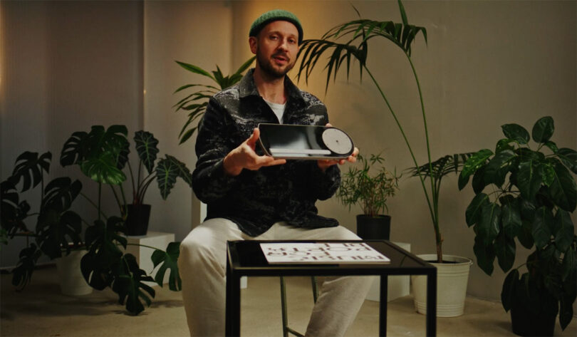 Designer Benjamin Großjohann holding his creation, the Coturn CT-1 turntable in front of small side table wearing green beanie cap and surrounded by house plants.