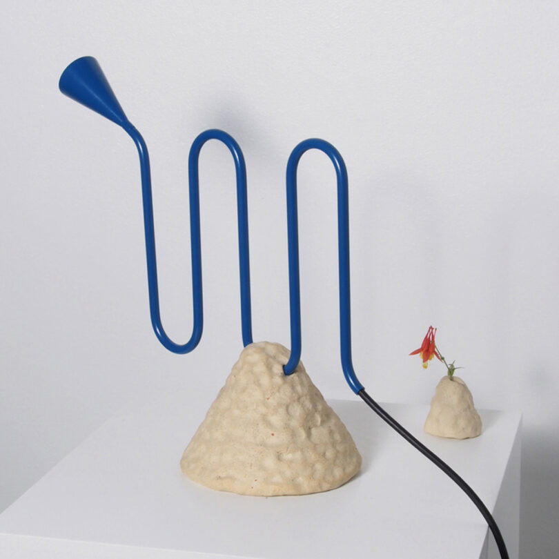 Blue LED table lamp with a wiggly wavy arm evocative of a brass instrument, paired with a ceramic cone base and black power cord set on a pedestal near a small flower.