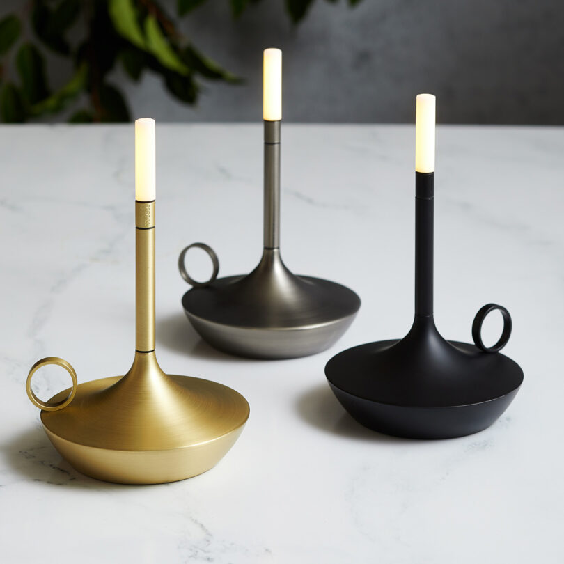 Three LED tipped candleholder shaped portable lights on a marble countertop – from left to right – brass, graphite and black. Each light tip is on a low light setting.